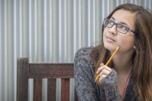 Pretty Young Daydreaming Female Student With Pencil Sitting on Bench Looking to the Side.